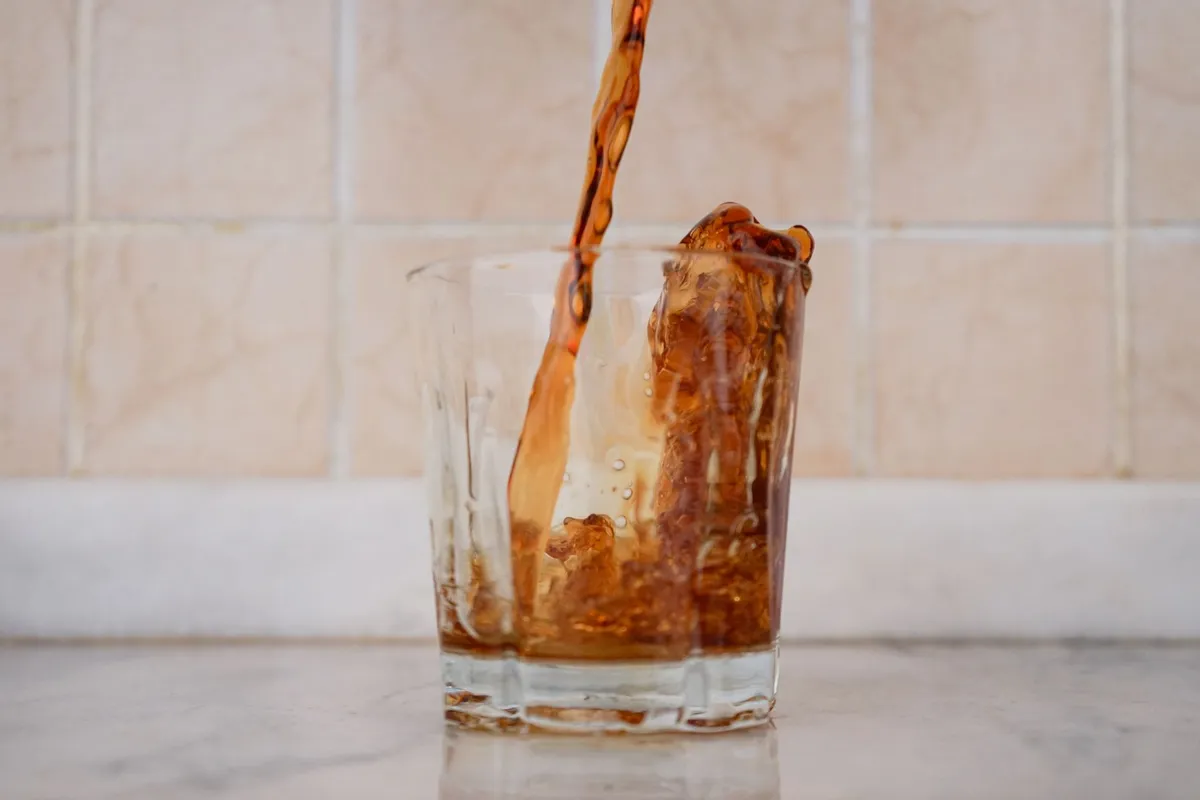 Carbonated soft drink being poured over ice cubes in a clear glass in natural light